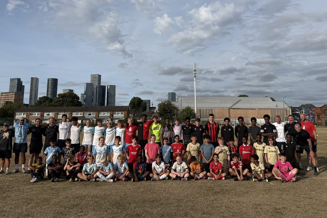 Old Trafford Juniors players and coaches lined up together on their home pitch in Manchester, with the city skyline behind them.