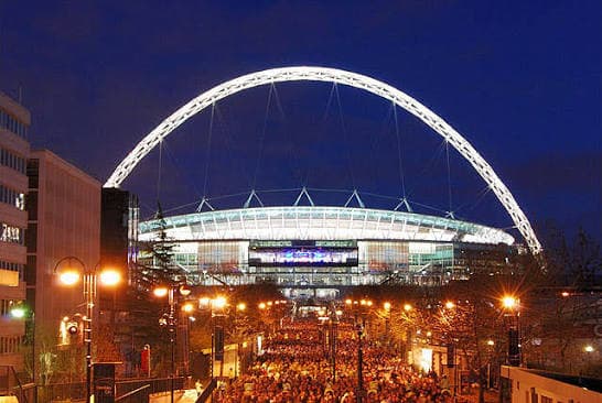 Wembley Stadium lit up at night with crowds walking along Wembley Way.